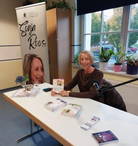 Autorin Sonja Roos sitzt bei einer Lesung an einem Tisch mit ihren Büchern vor einem Roll-up.