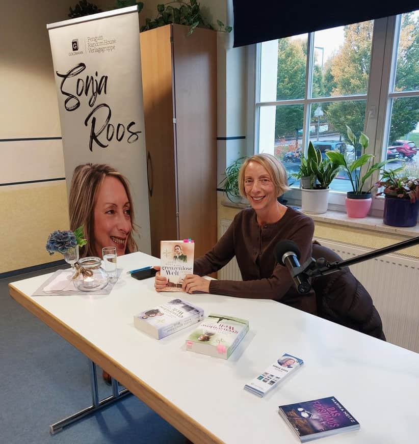 Autorin Sonja Roos sitzt bei einer Lesung an einem Tisch mit ihren Büchern vor einem Roll-up.